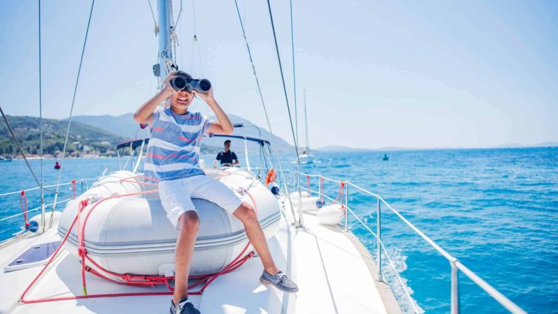 Cute boy with binoculars on board of sailing yacht on summer cruise. Travel adventure, yachting with child on family vacation. Kid clothing in sailor style, nautical fashion.